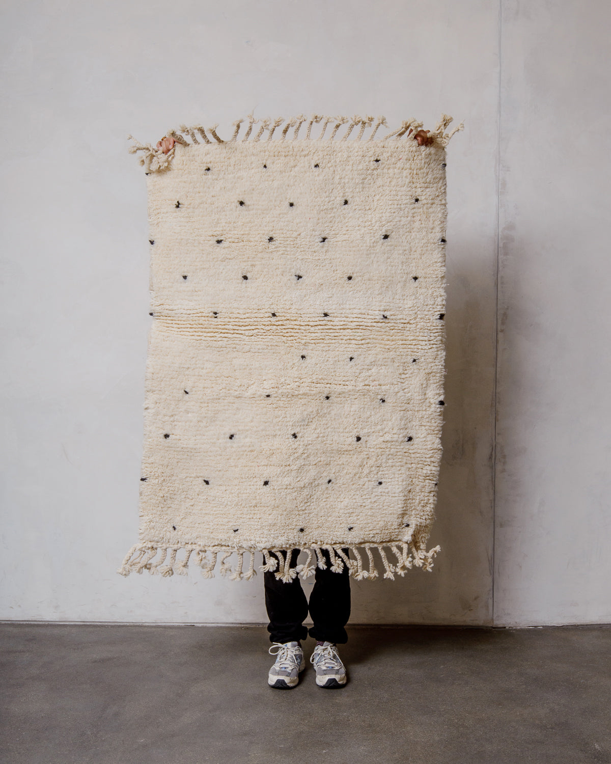 Small cream Berber rug with black dots being held up by a person in front of a white wall.